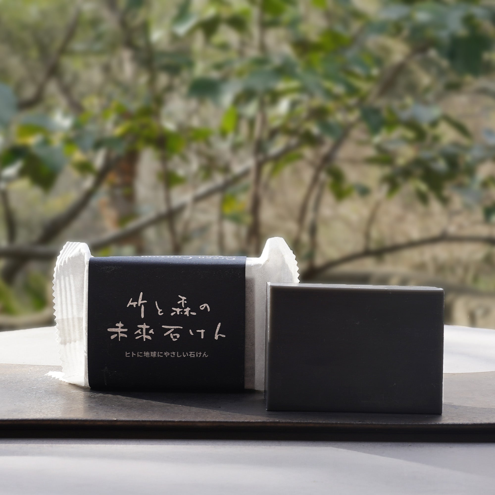 緑豊かな風景を背景にした、包装済みの石けんと本体。ひとつひとつ丁寧に作られた、手仕事の温もりが伝わる製品の佇まい